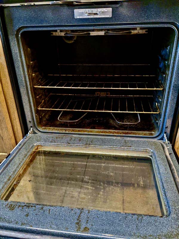 An open, empty oven with two interior wire racks and a drop-down door. The interior is visibly worn and stained, suggesting it has been used frequently. The oven's control panel is not visible in the image.