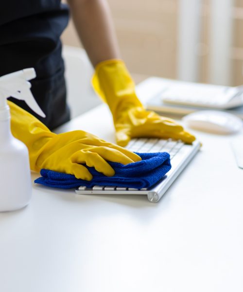 A person wearing yellow rubber gloves cleans a computer keyboard with a blue cloth. A white spray bottle is on the desk beside the keyboard, and a computer mouse and monitor are visible in the background.